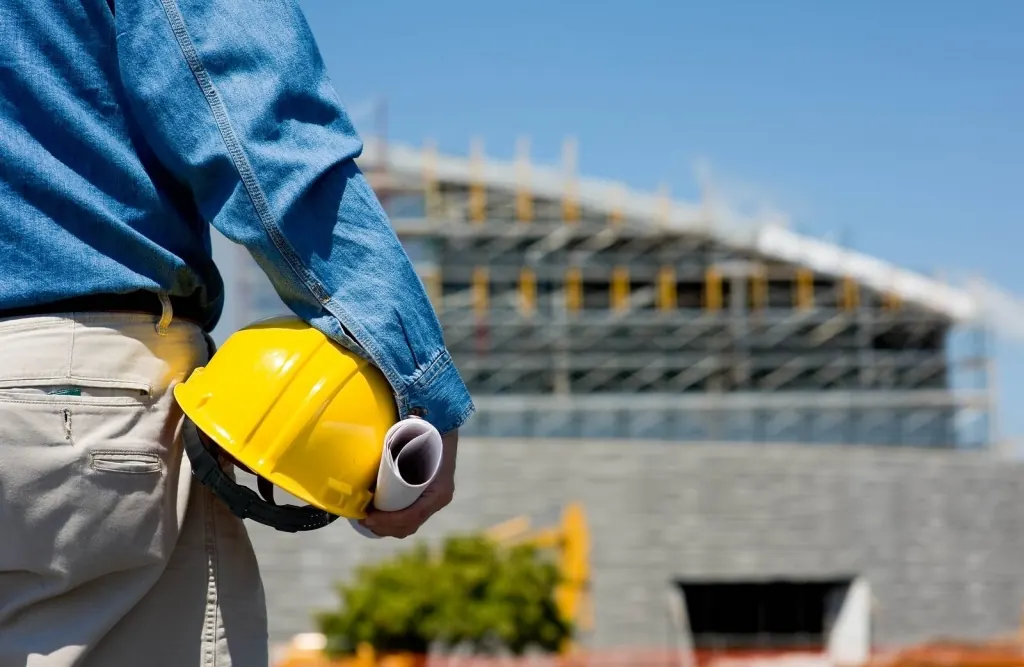 Profissional com capacete e planta enrolada em frente a obra em estrutura metálica