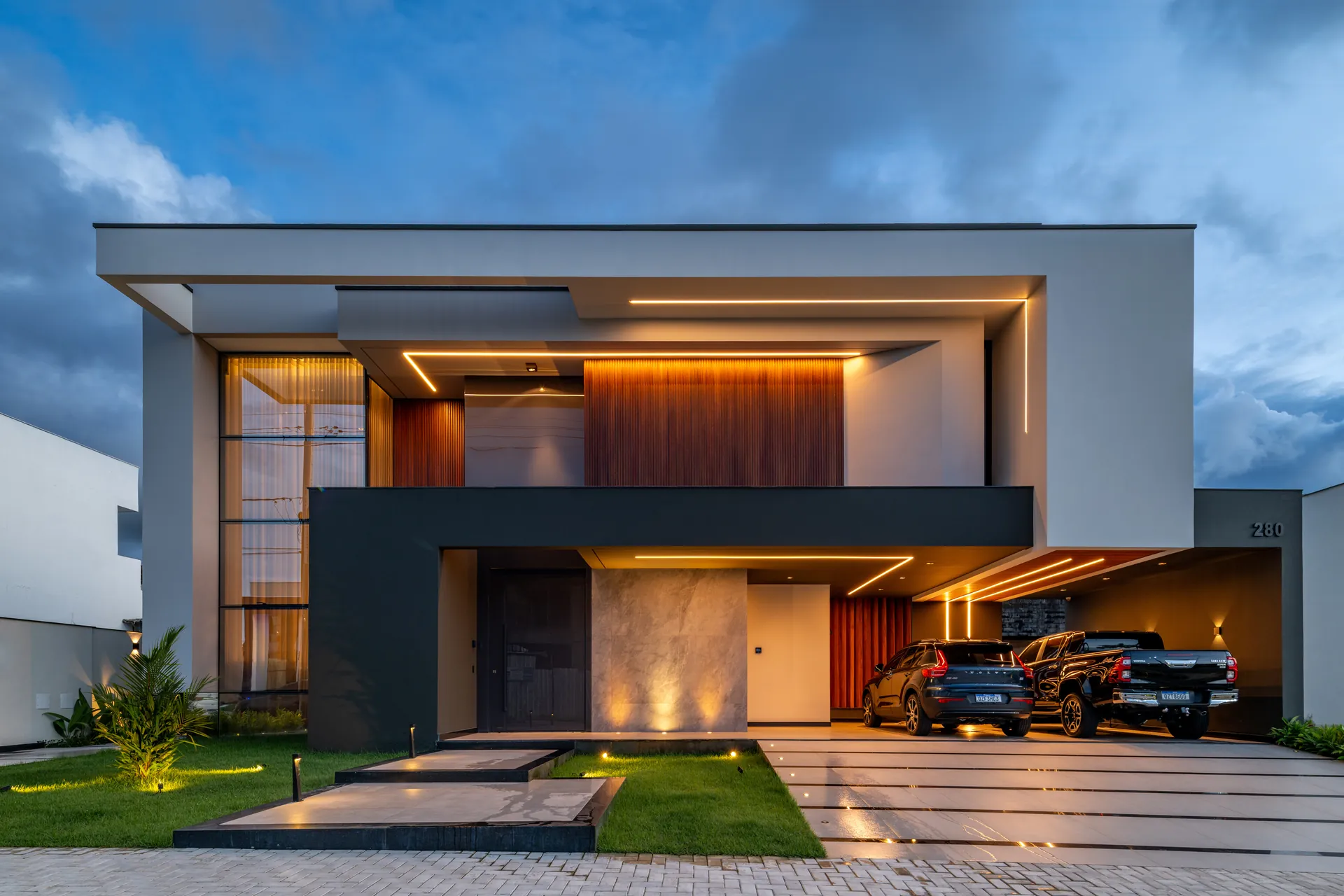 Fachada da Residência L|D ao entardecer, com volumes retos, madeira e iluminação na garagem